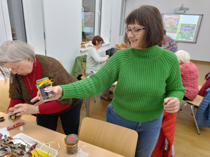 Uśmiechnięta seniorka w zielonym sweterku i okularach prezentuje swoją kompozycję w słoiku, która nazwała "Dary jesieni". W słoiku znajdują się suszone owoce i liście. Wieczko przyozdobione jest zielona bibułą i przewiązane żółtą wstążką.