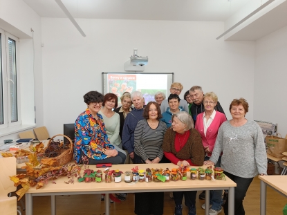 Grupa uśmiechniętych seniorów i seniorek pozuje do zdjęcia z prowadzącą zajęcia "Jesień w słoiku". Przed niemi na stole prezentowane są słoiczki z jesiennymi kompozycjami własnoręcznie przygotowanymi przez seniorów. 