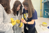 Nauczycielka z granatowej koszulce i długimi włosami związanymi w kitkę pokazuje dwóm uczennicom ubranym w białe kitle  i żółte rękawiczki gumowe, jak obsługiwać pipetę.