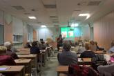 Seniorzy siedzą przy ławkach sali ćwiczeniowej. Prowadząca pokazuje owoce, na których rozwijają się choroby grzybowe.
