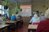 Seniorzy siedzą przy ławkach w sali ćwiczeniowej, prowadząca przedstawia informacje dotyczące rozmnażania roślin, w tle na ekranie pokazywana jest prezentacja multimedialna, z sufitu zwisają rośliny w formie kokedamy - wiszącego ogrodu.
