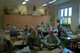 Seniorzy obwiązują sznurkiem bryłę korzeniową rośliny wraz z ziemią i osłoniętą mchem. Seniorzy znajdują się w sali ćwiczeniowej, siedzą przy ławkach.