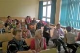 Siedemnaścioro seniorów i seniorek siedzi przy ławkach w sali ćwiczeniowej i słucha wykładu.
